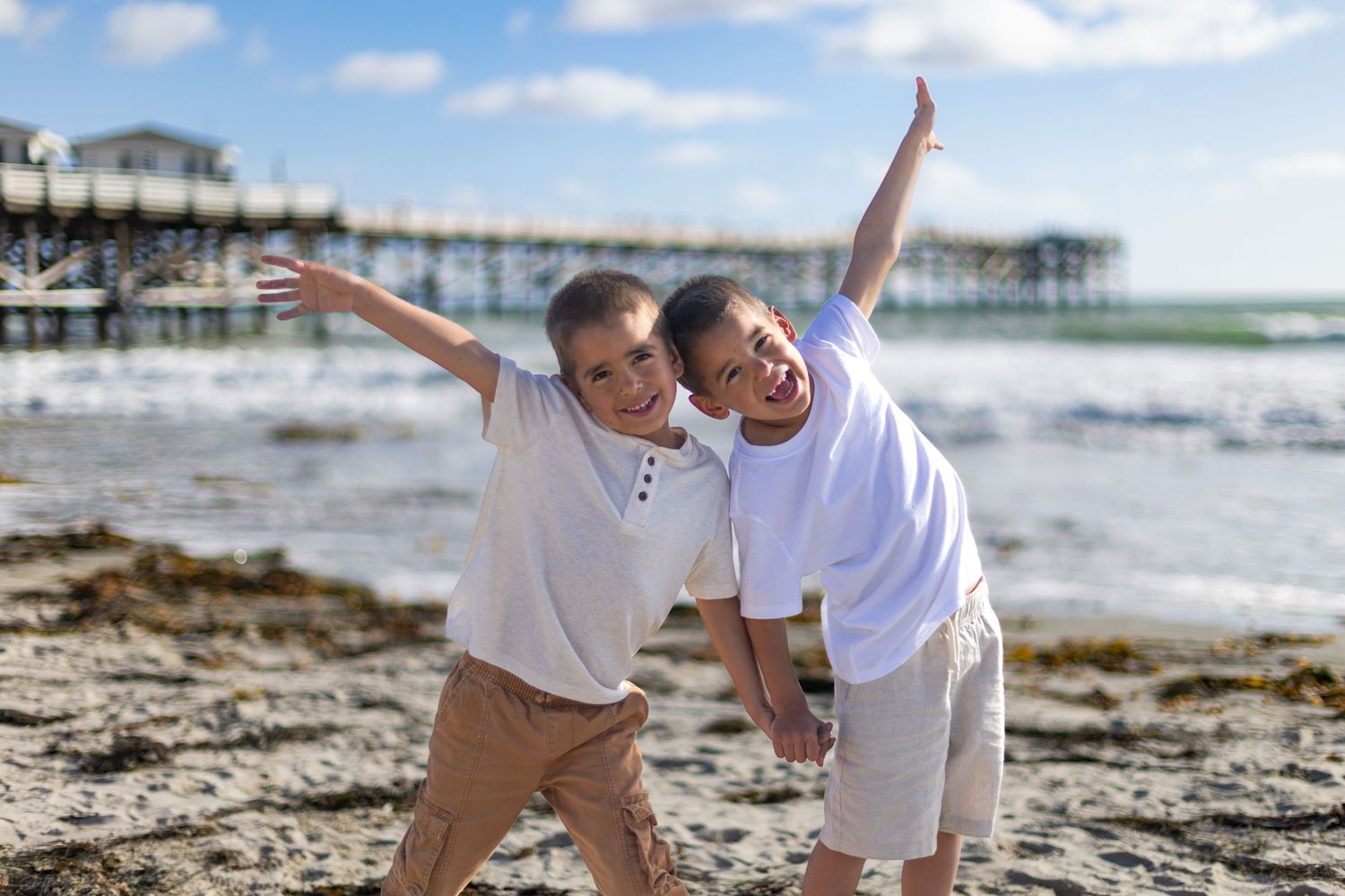 San Diego family photography of twin boys posing playfully on the beach with the ocean and a wooden pier in the background.