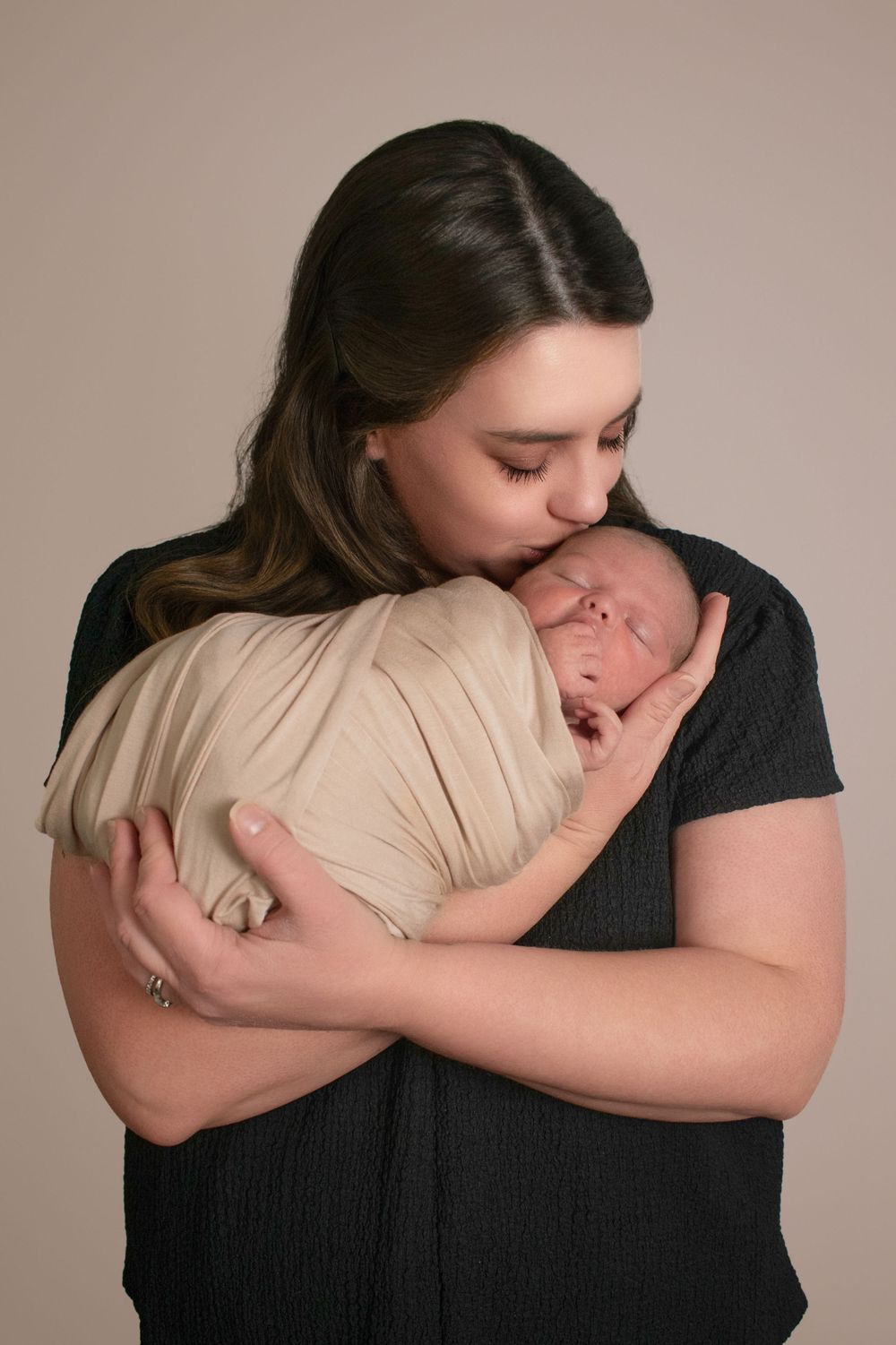 A mother in a black shirt tenderly kisses her swaddled newborn baby wrapped in beige cloth against neutral background baby photos SC