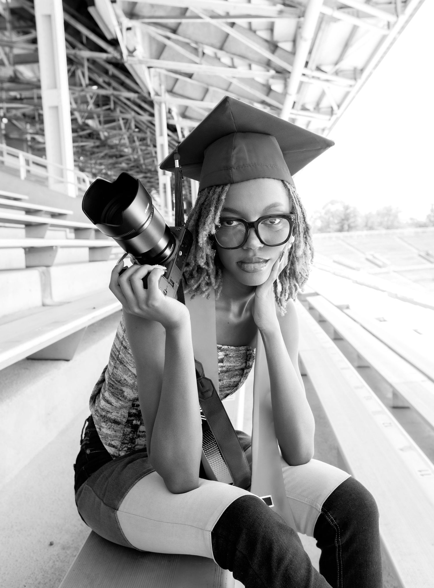 Graduate in cap and gown poses with camera in black and white photo.