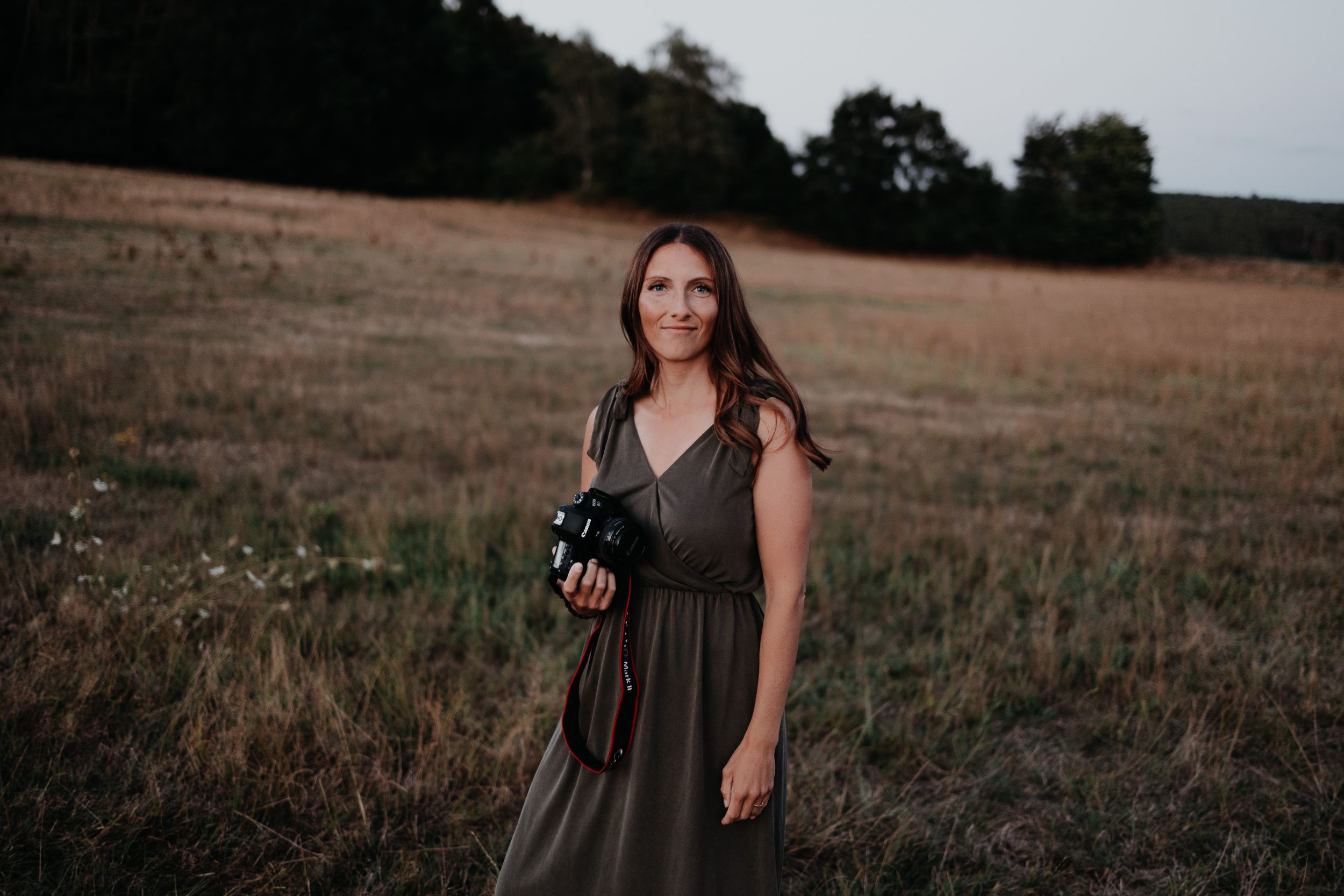 A woman in a brown dress stands in a dusky field with trees in the background.