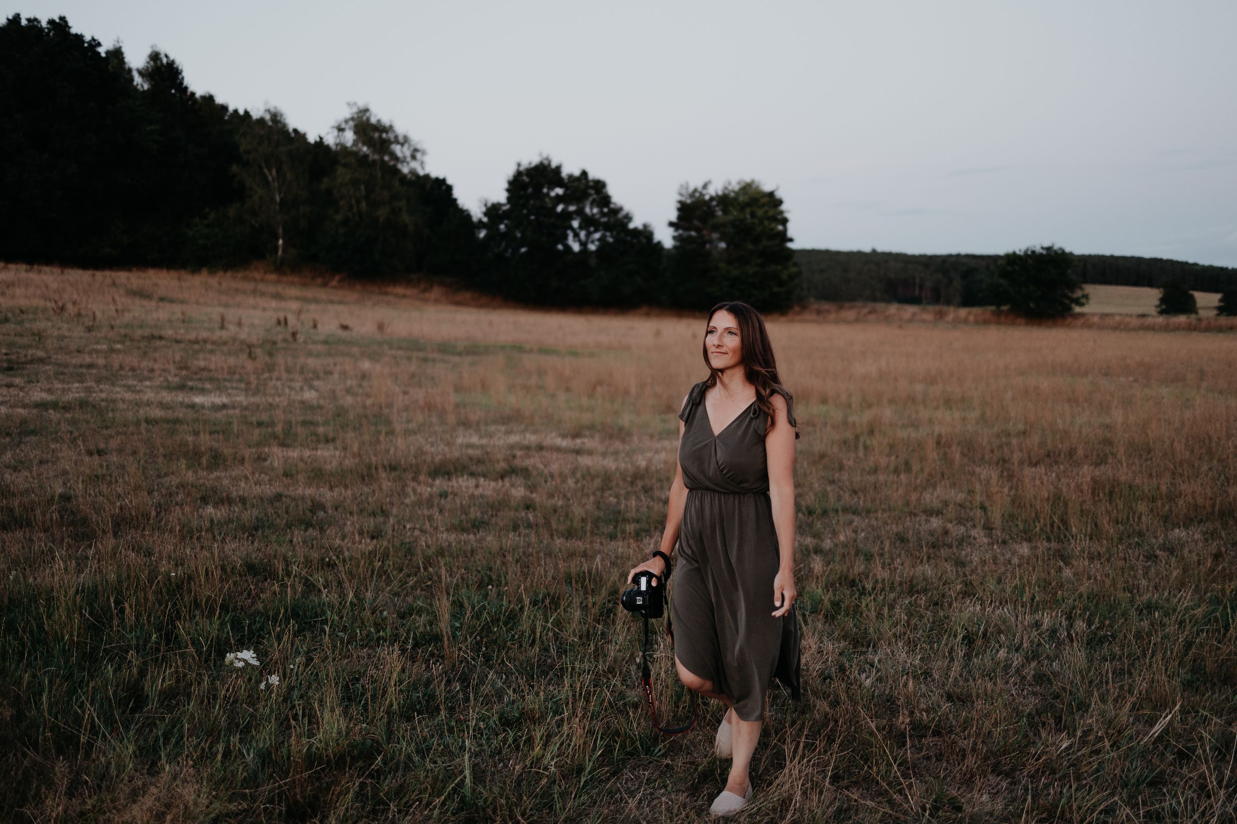 A person in a brown dress walks through a golden field at dusk with trees in the background.