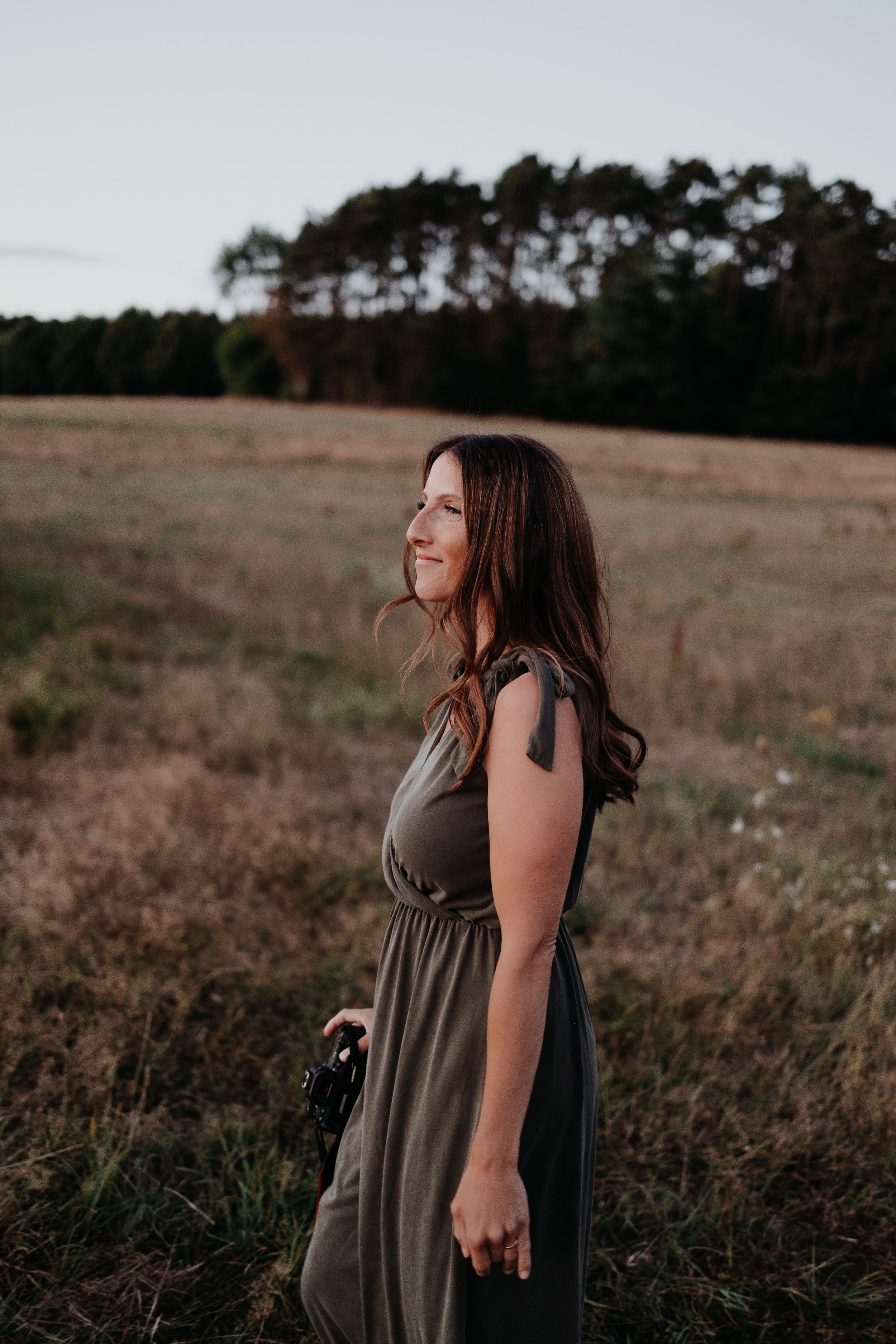 Person in a flowing olive dress laughs joyfully while standing in a rural field at sunset with trees in background.