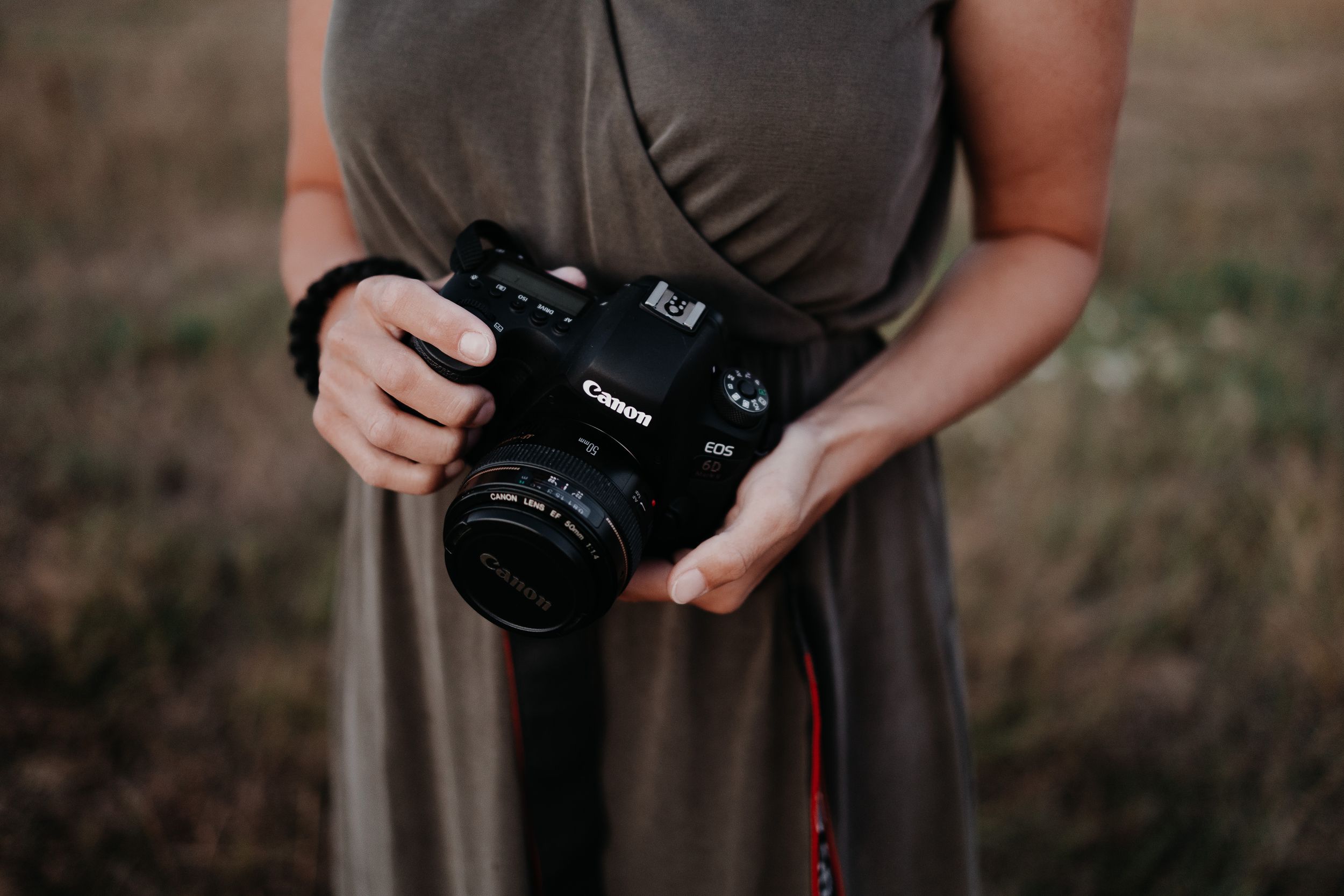 Person in a brown dress holding a black Canon DSLR camera with lens against a blurred outdoor background.