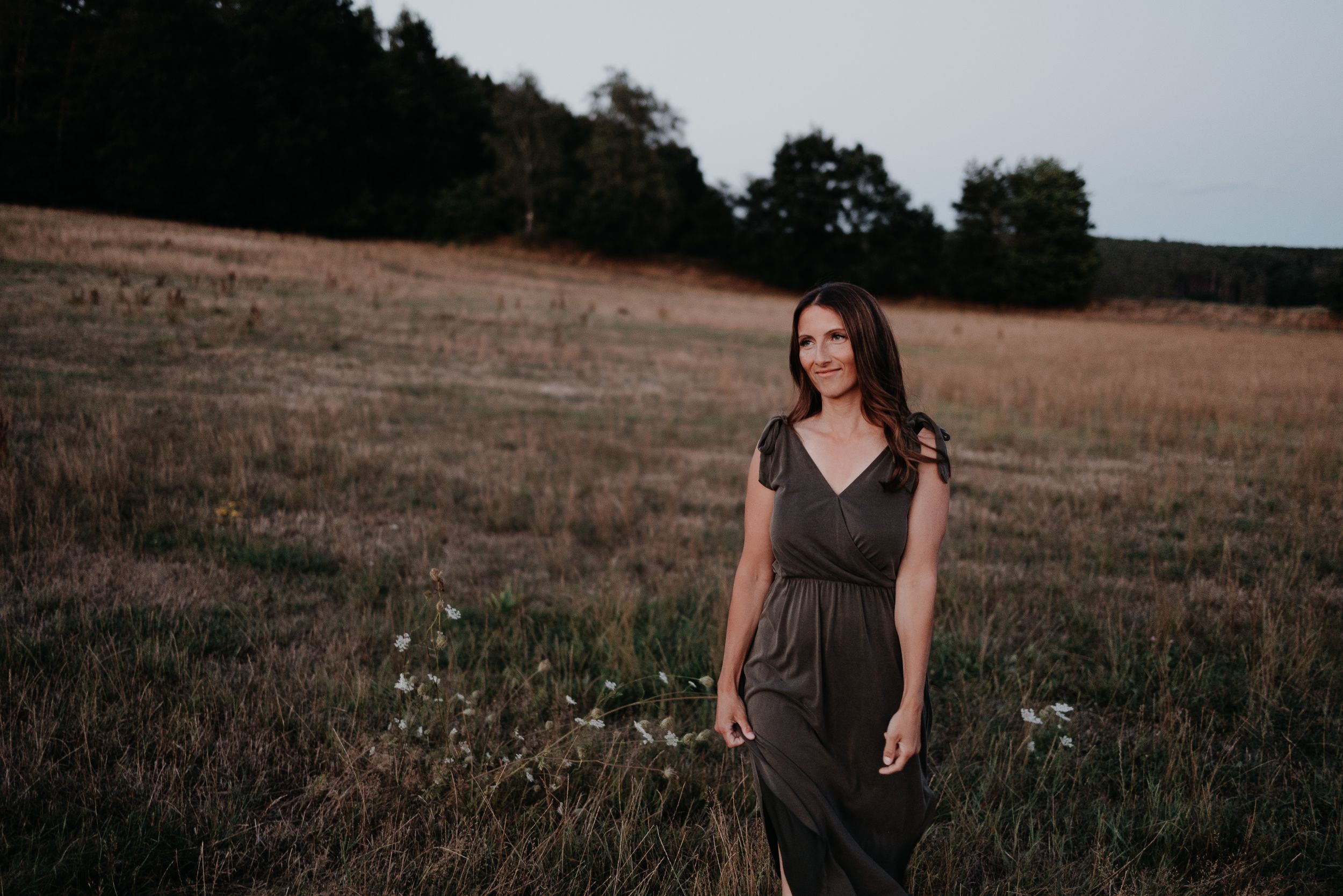 Someone wearing a brown dress stands in a golden field at dusk with trees in the background.