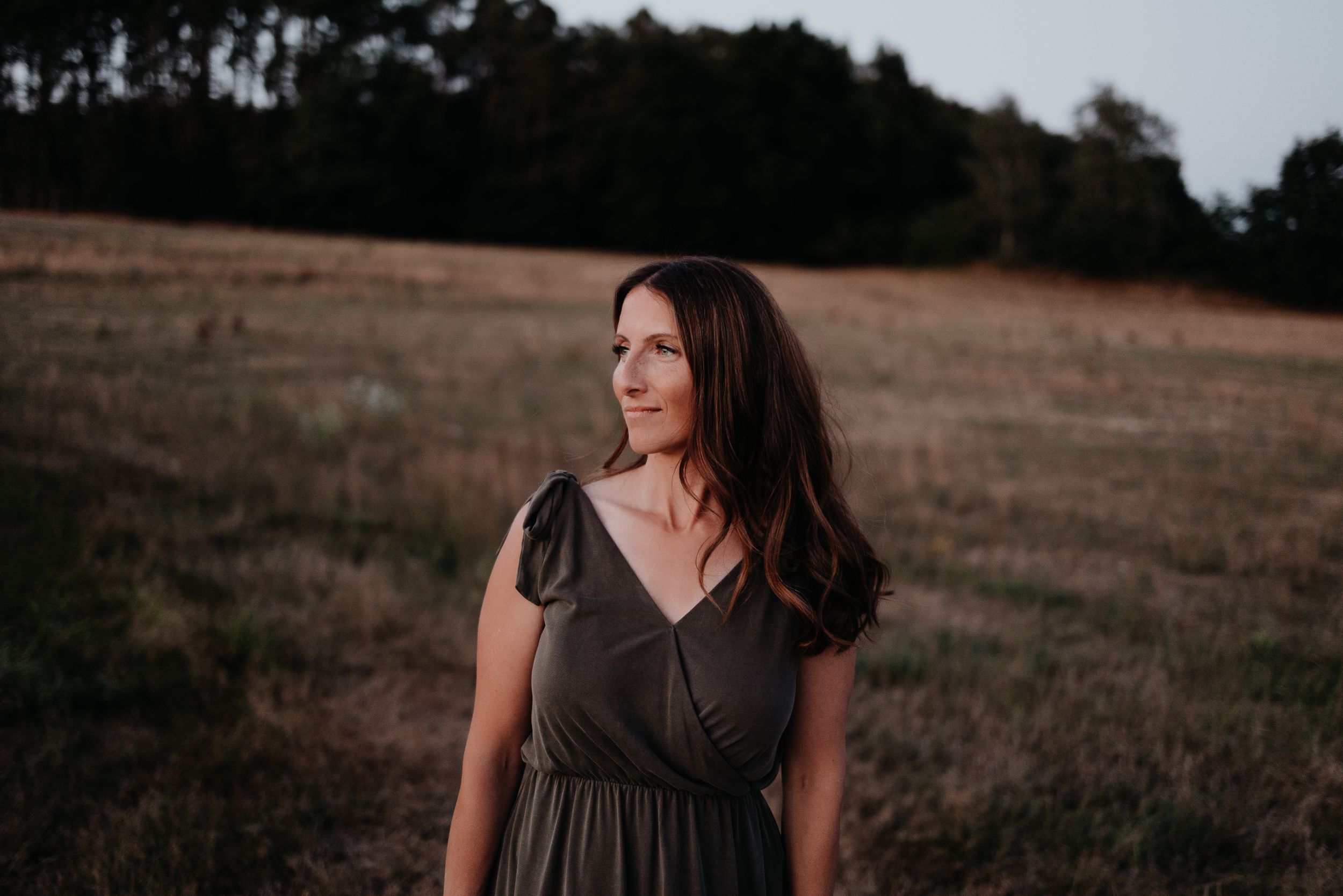 Long brown hair and gray dress standing in open field during sunset with trees in background.