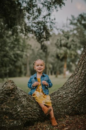 A child sits playfully on a large tree branch in a park wearing a denim jacket and yellow outfit.