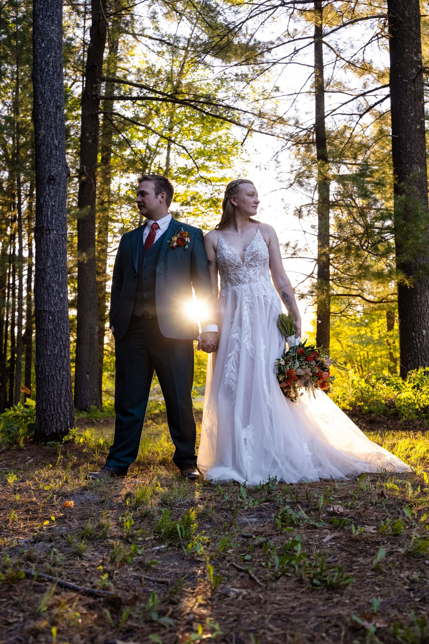 A couple in wedding attire poses in the woods at sunset with sunlight glowing through tall evergreen trees.