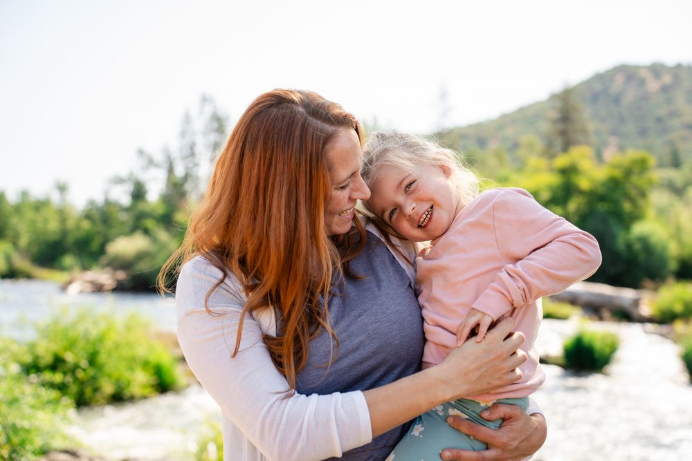 Mom cuddles and laughs in a portrait with her daughter by the river