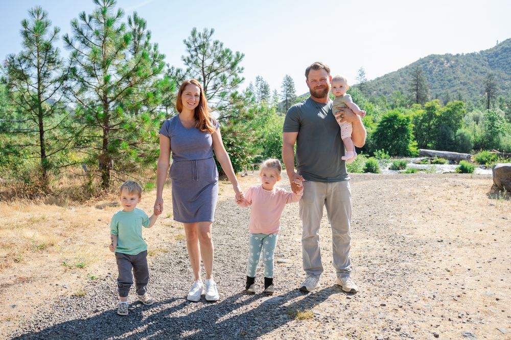 Mom and dad walk with 2 toddlers and a baby girl in a family portrait by the trees and Rogue River