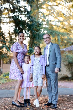 Smiling family of four standing outdoors at golden hour, parents with two daughters in purple and floral dresses.