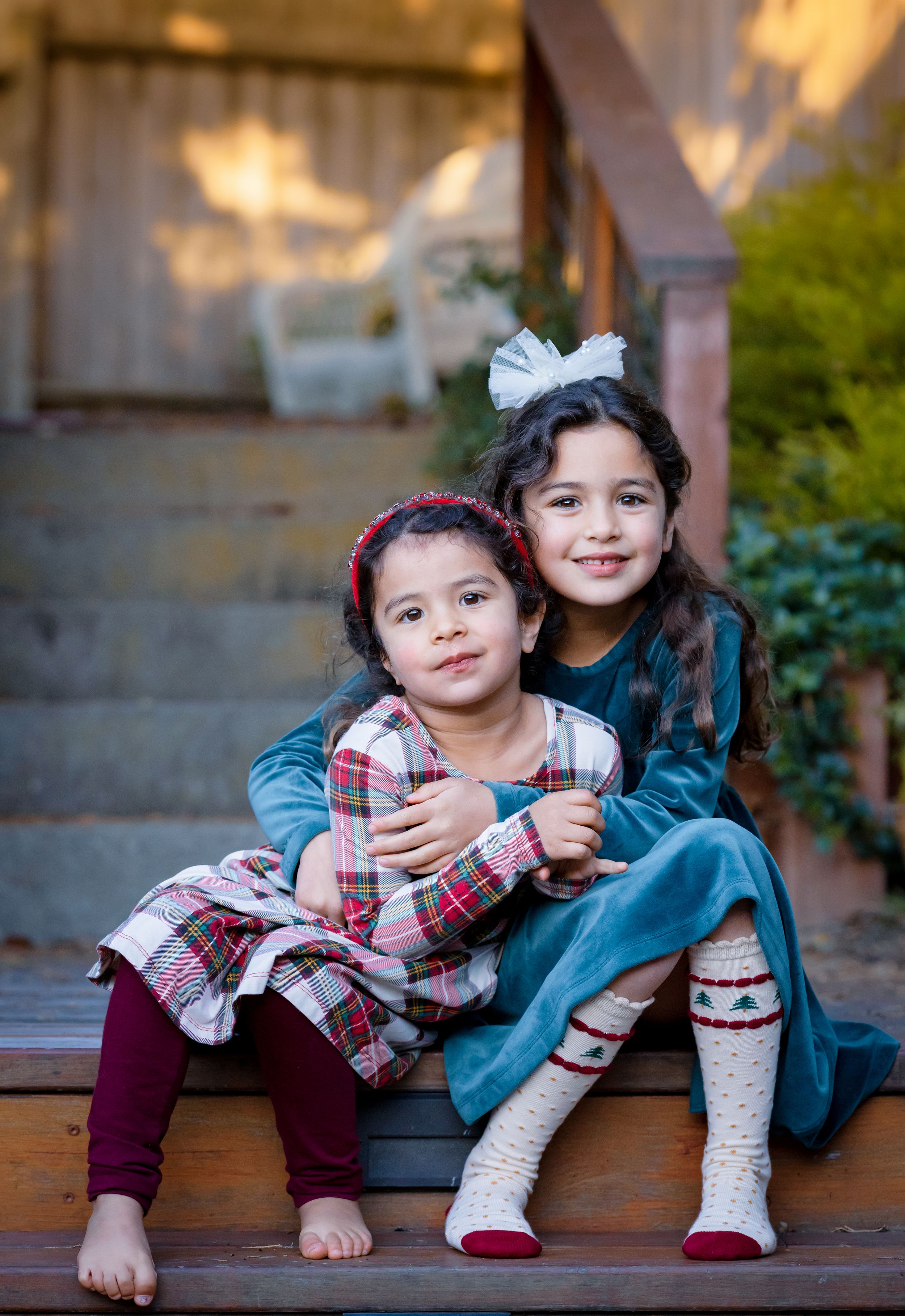 Two young sisters in holiday outfits sit together on wooden steps, older sister hugging younger sibling.