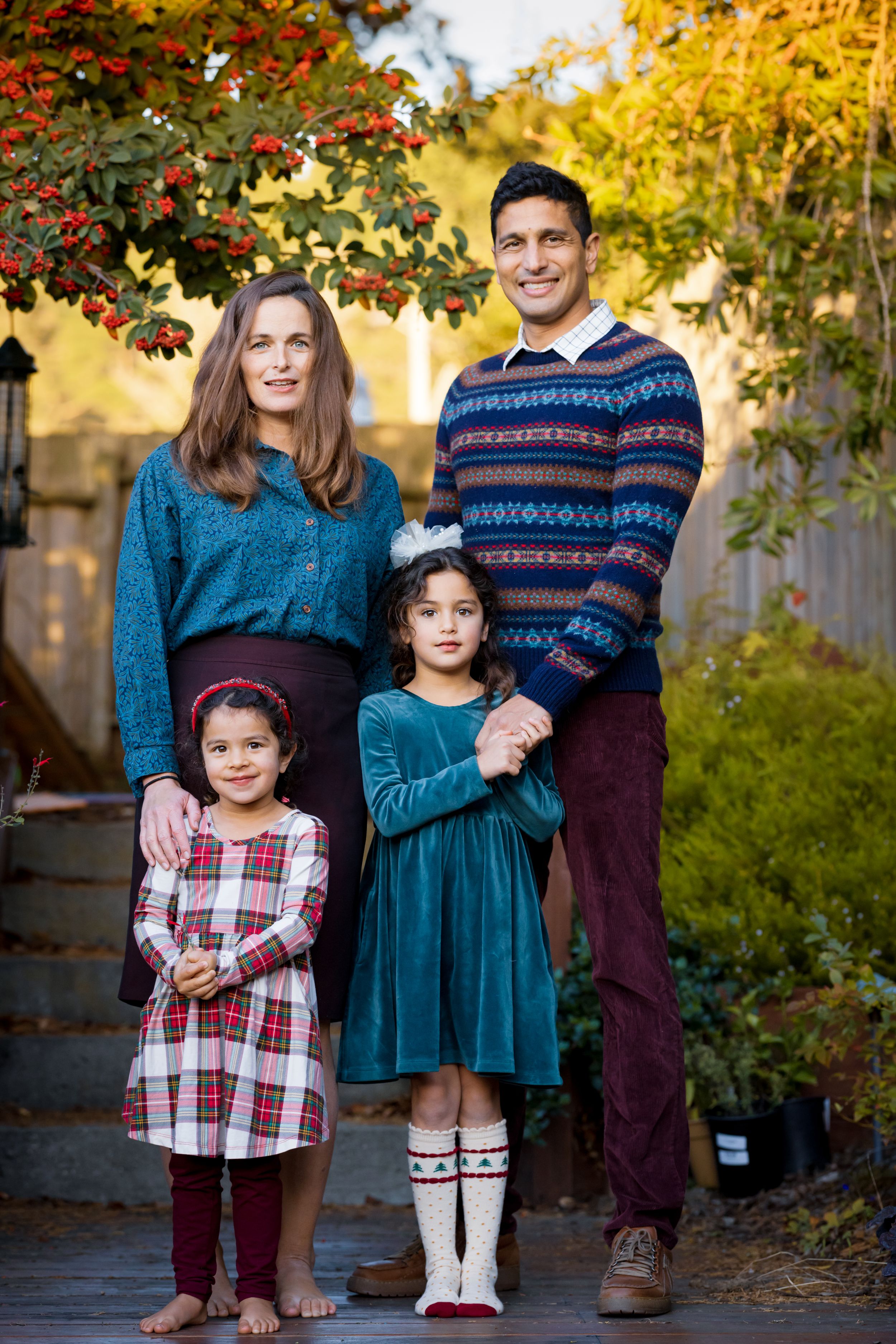 Family of four posing outdoors in autumn garden, parents with two young daughters in coordinating teal and burgundy outfits.