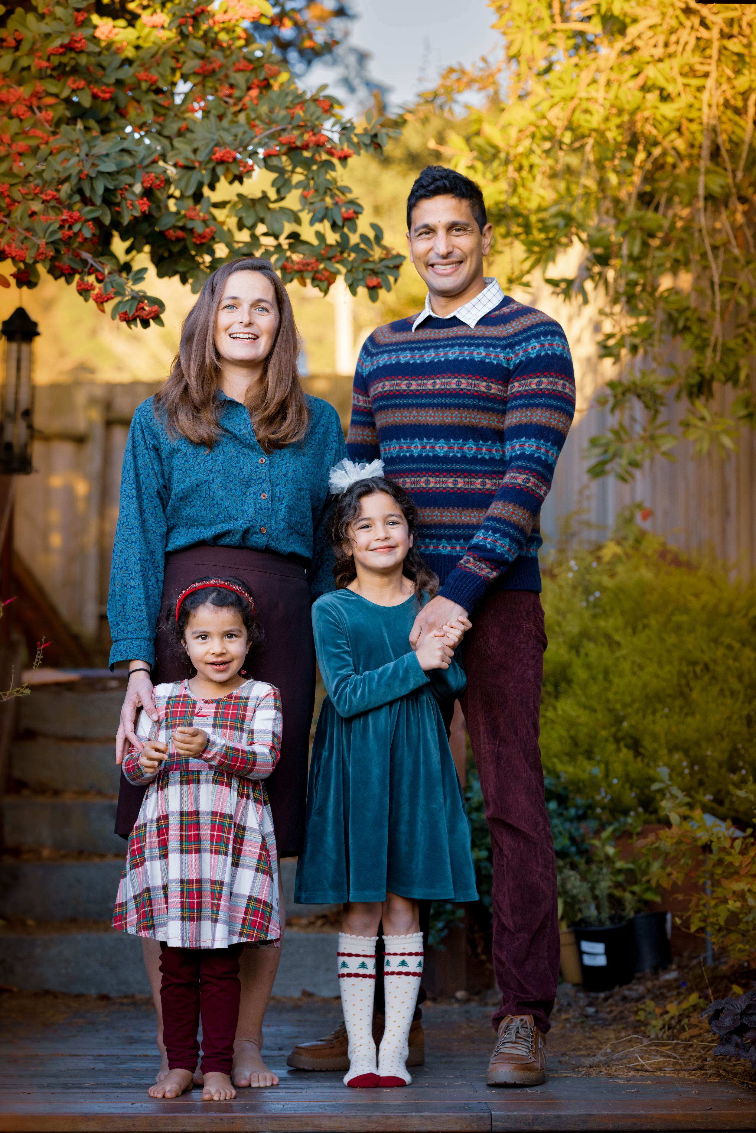 Smiling family portrait with parents and two girls in matching teal and plaid outfits surrounded by fall foliage.