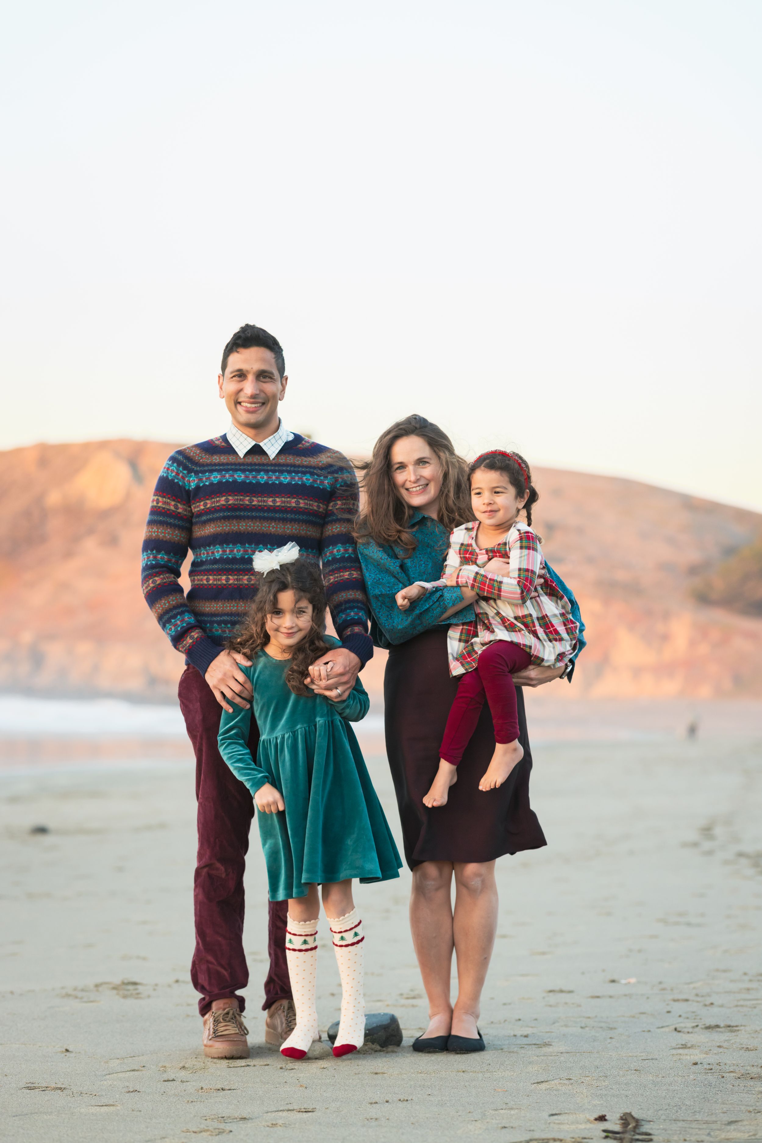Happy family of four poses on a sandy beach at sunset, wearing coordinated fall holiday outfits.