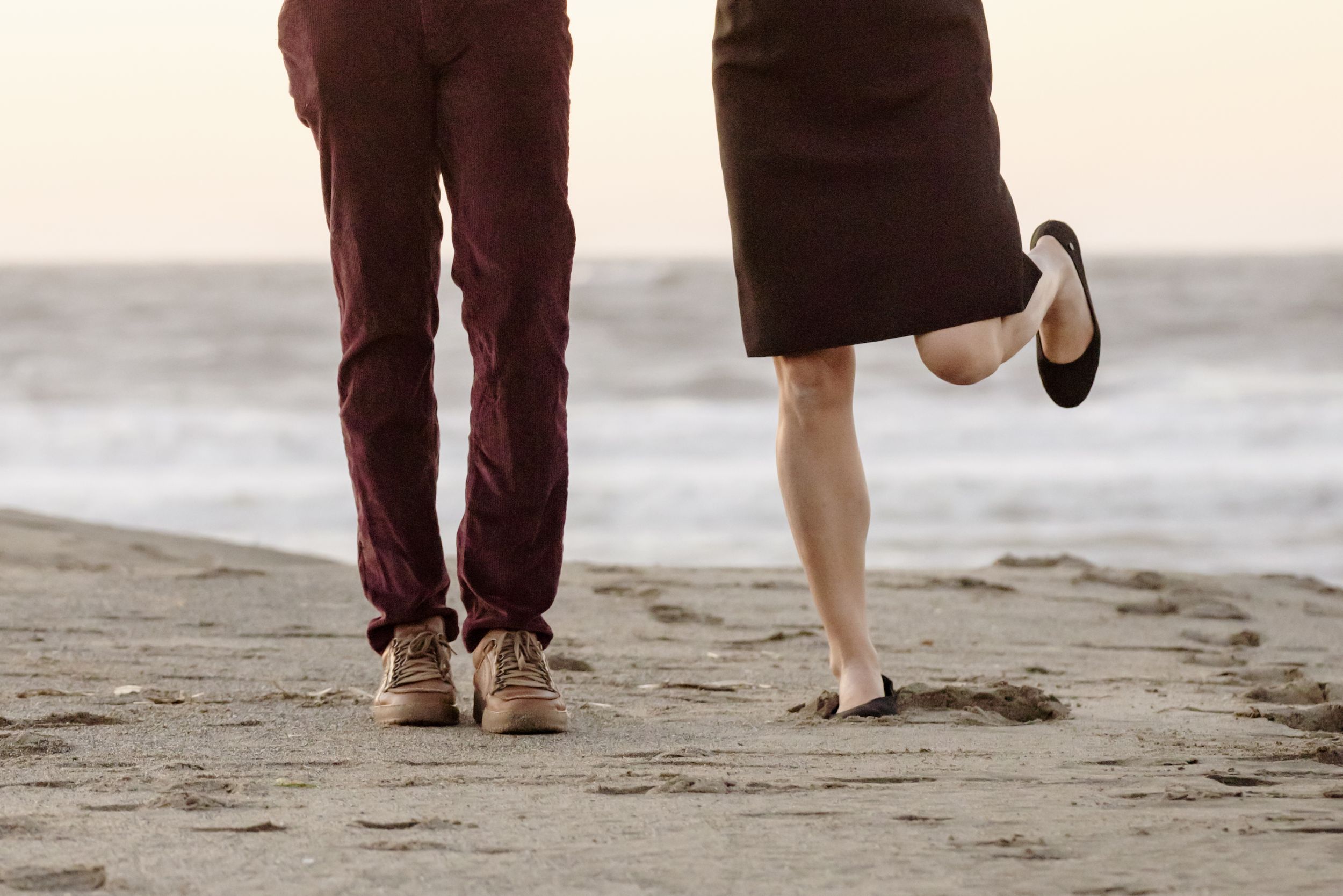 Couple standing on a sandy beach at sunset, man in burgundy pants and woman in dark skirt, barefoot.