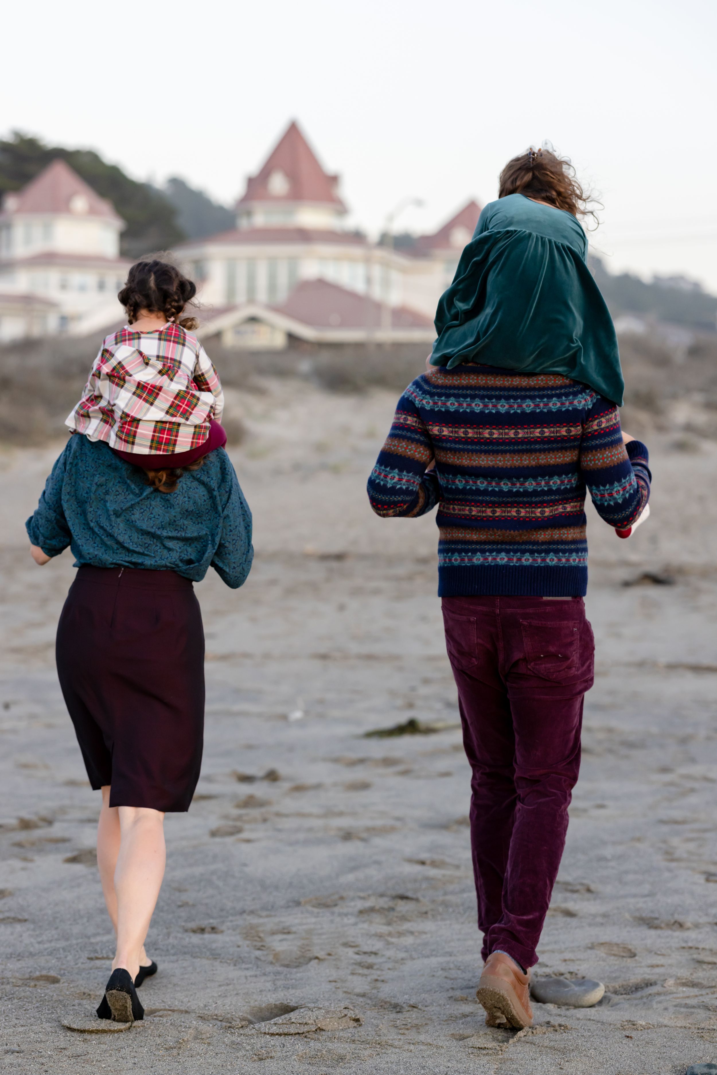 Parents carrying children on their shoulders walk away on a beach toward a historic building at dusk.