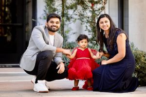 Young South Asian family with toddler girl in red dress posing outside modern building entrance.