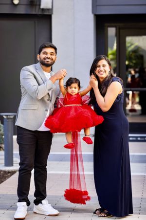 Joyful South Asian parents lifting toddler daughter in red tulle dress outside modern building.