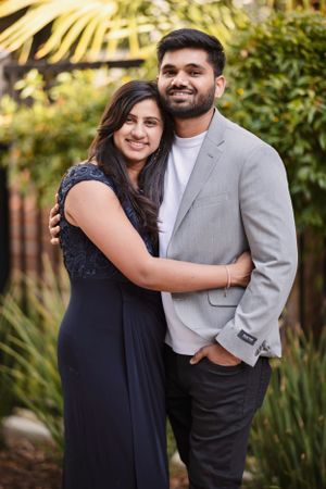Smiling South Asian couple embracing outdoors, woman in navy dress and man in gray blazer with white shirt.