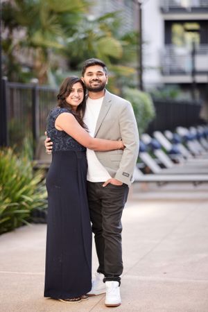 Happy South Asian couple posing together outdoors, woman in navy gown hugging man in gray blazer and white sneakers.