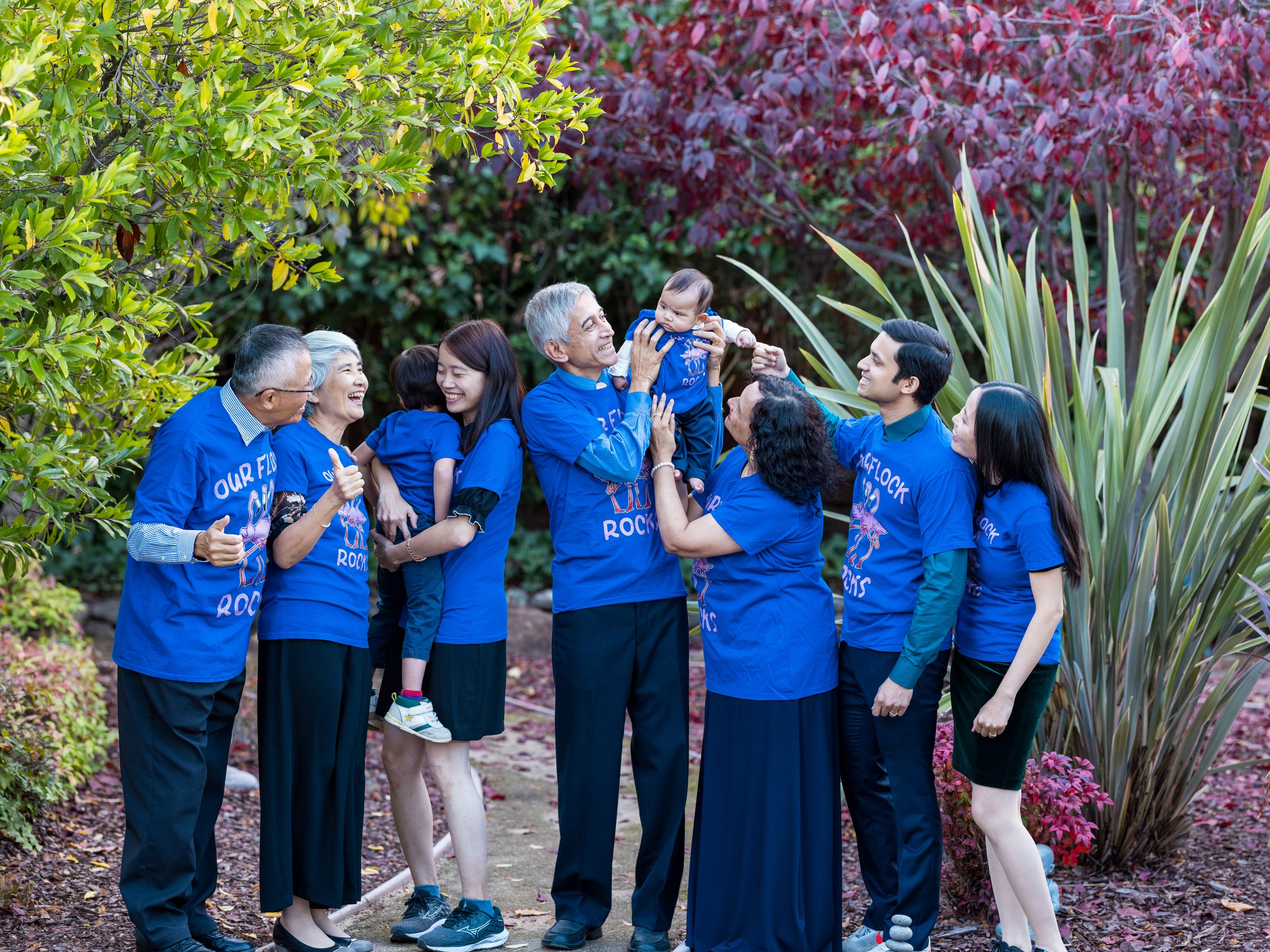 A joyful multigenerational Asian family in matching blue shirts gathers outdoors among colorful garden foliage.