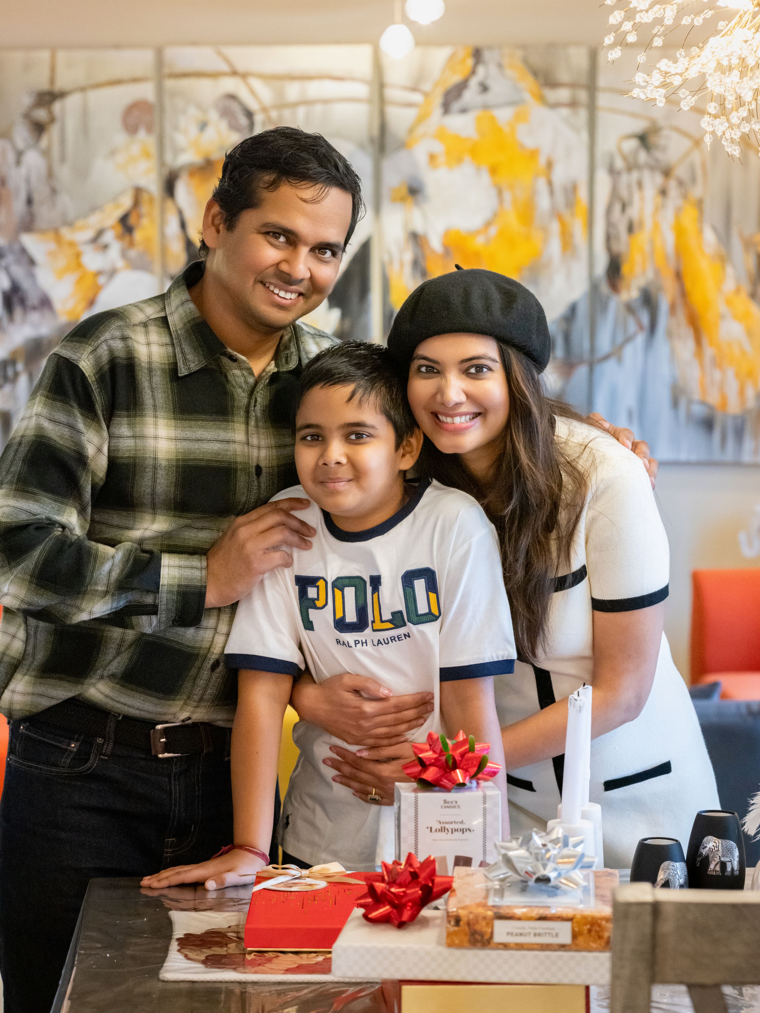 South Asian family of three smiling together indoors with gifts and decorative artwork in background.