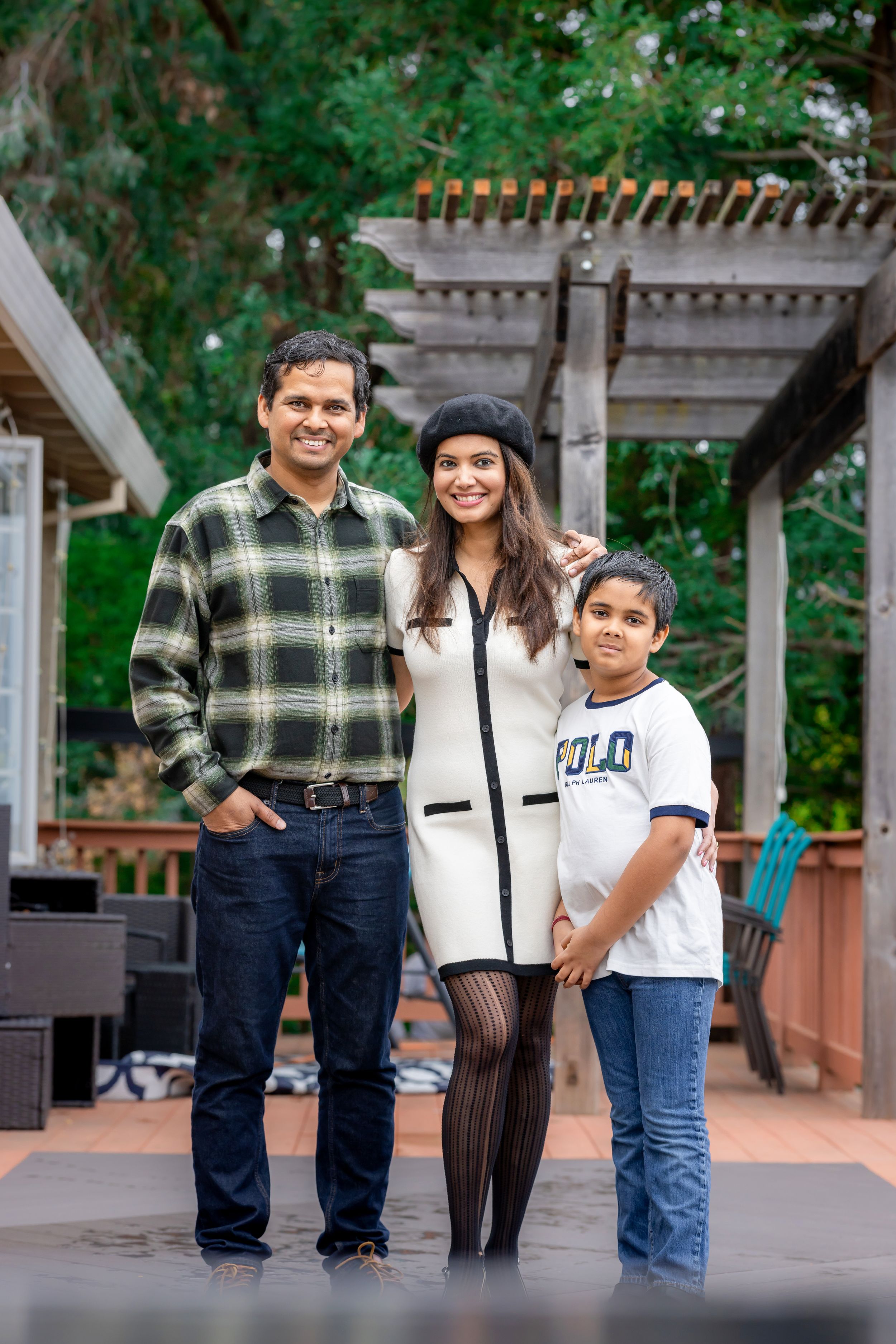 South Asian family of three smiling together on outdoor patio with pergola and lush trees behind.