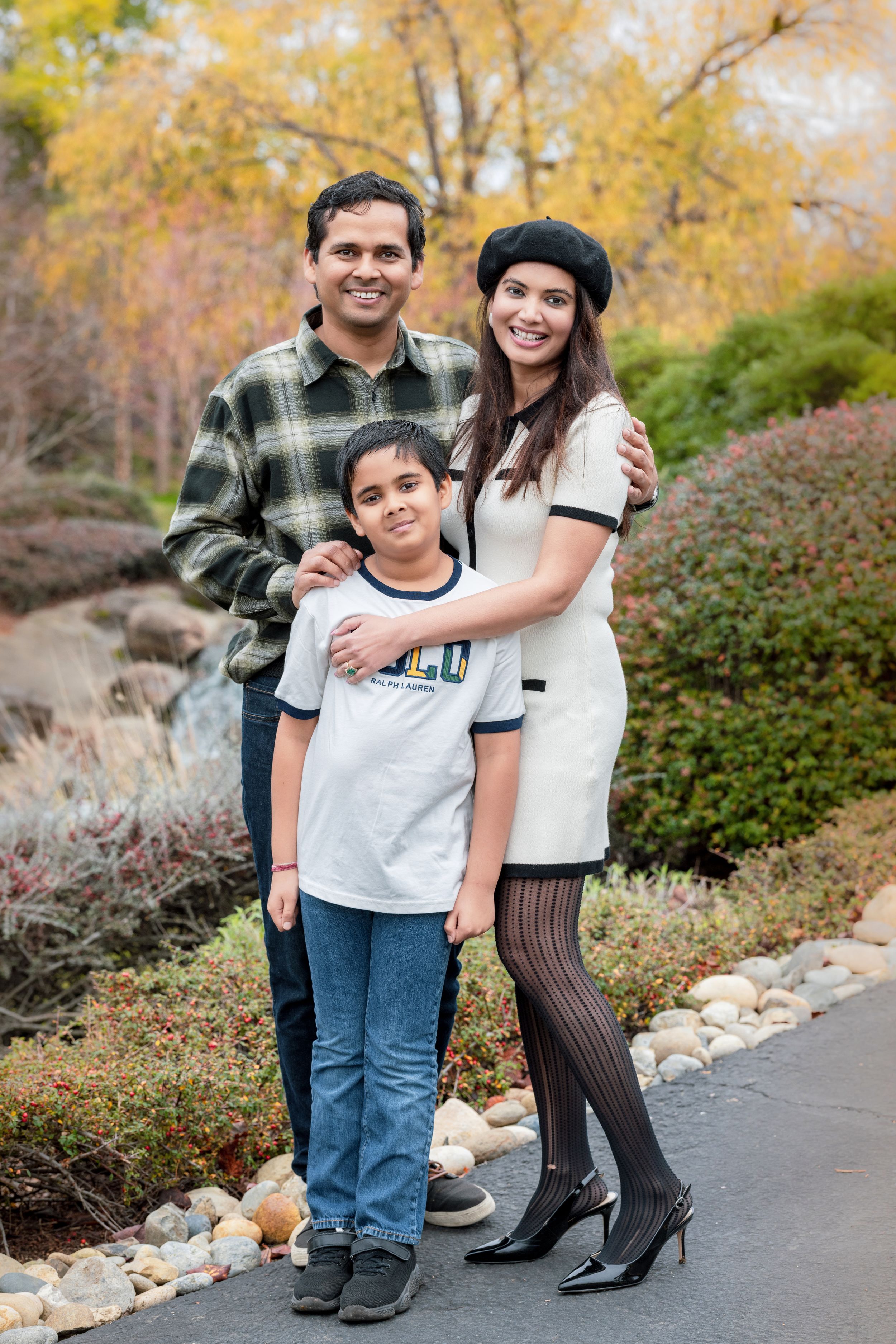 South Asian family of three posing outdoors in autumn garden with colorful fall foliage backdrop.