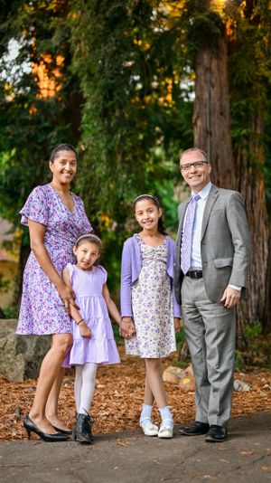 Family of four posing outdoors among tall trees, two girls in lavender dresses with parents dressed elegantly.