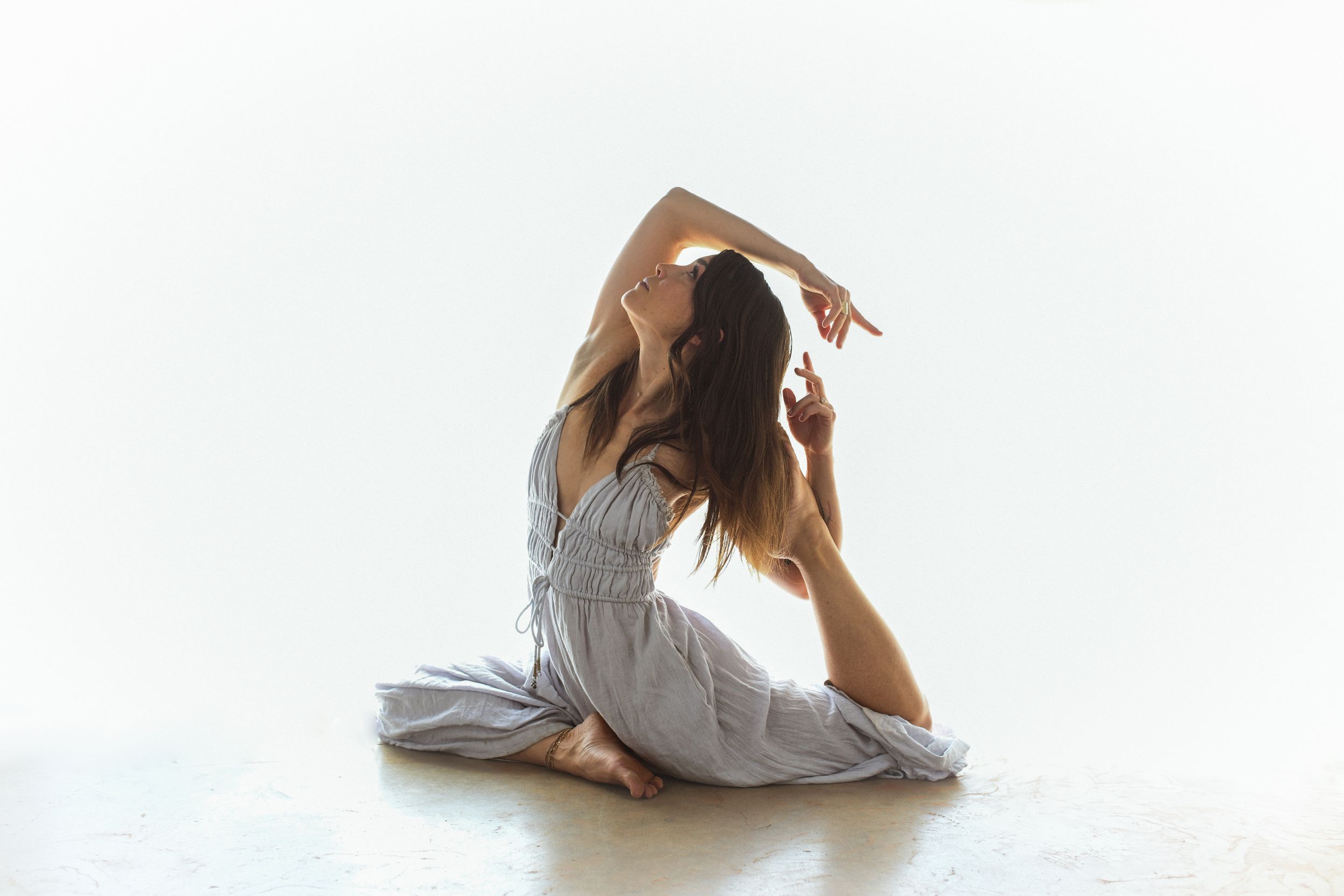 Graceful dancer in flowing dress performs artistic stretch pose on white background.