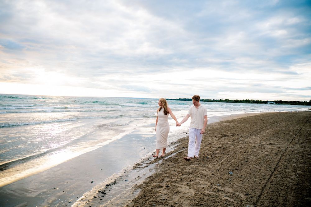 Couple in white outfits walking along a serene beach at sunset with waves crashing on the shore.