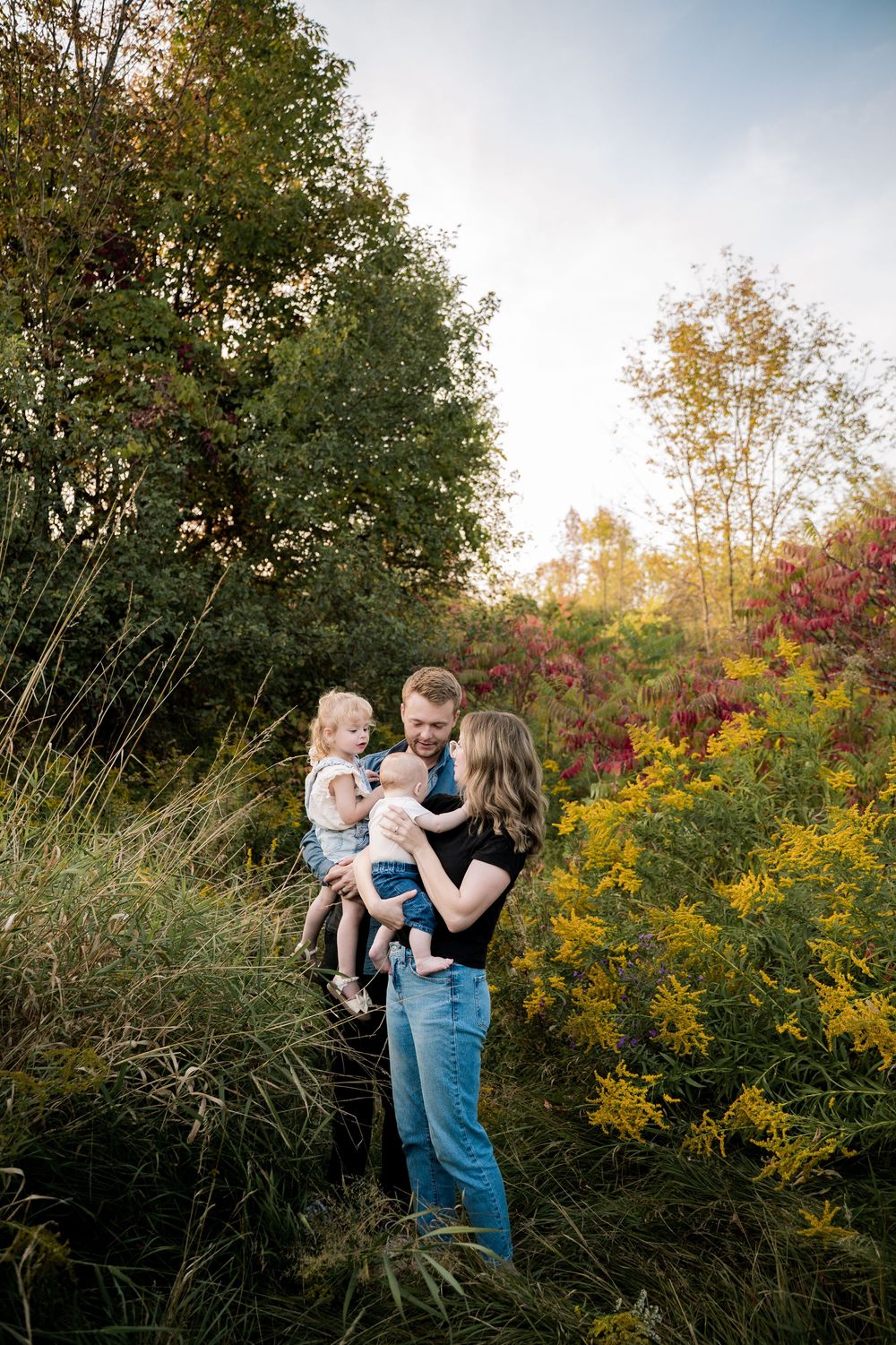 A family enjoys a tender moment together against a backdrop of autumn foliage and golden sunlight in a natural outdoor setting.