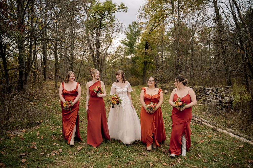 A bridal party wearing red dresses walks together through a wooded forest path during an autumn wedding.