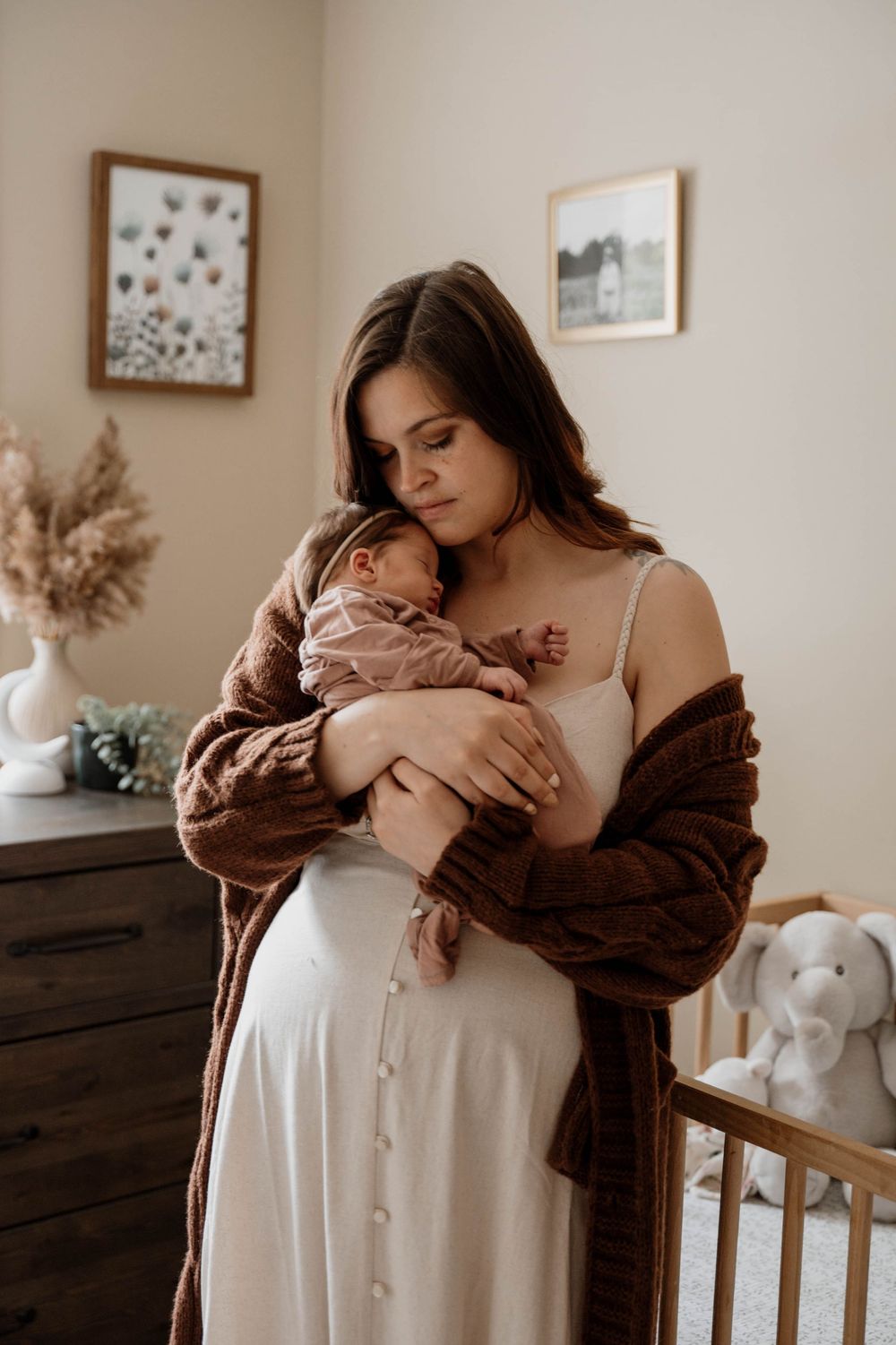 A person in a brown sweater embraces a baby in a white dress near a wooden crib in a neutral-toned nursery.
