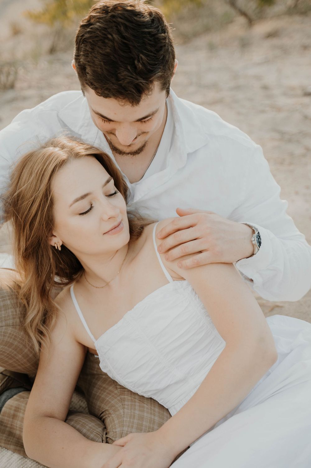 Intimate romantic couple in white clothing sharing a tender moment while reclining outdoors in soft sunlight.