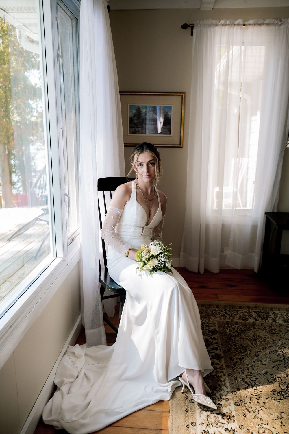 A person in an elegant white wedding dress sits gracefully by a sunlit window in a room with floral carpeting.