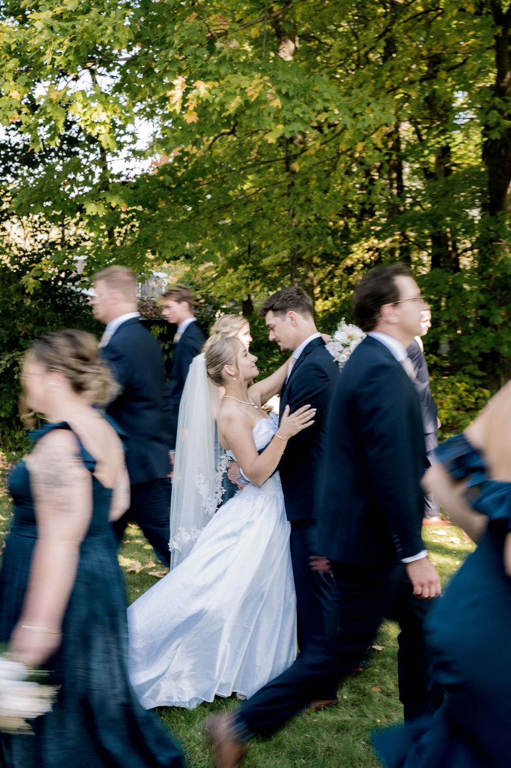 A wedding party in motion creates a blurred action sequence of people walking through a green outdoor setting.