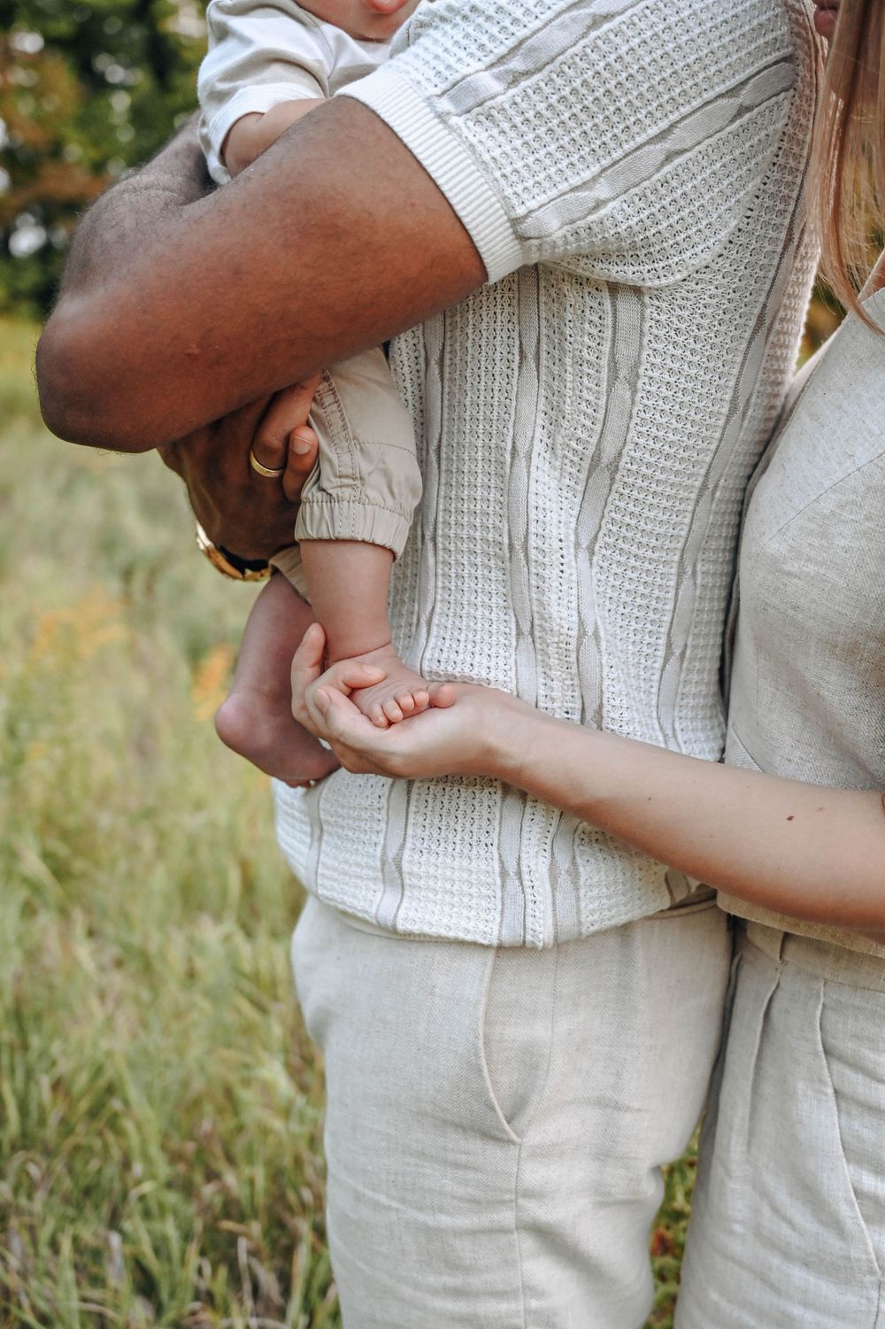 Close-up of couple holding small baby's feet in white knit clothing outdoors.
