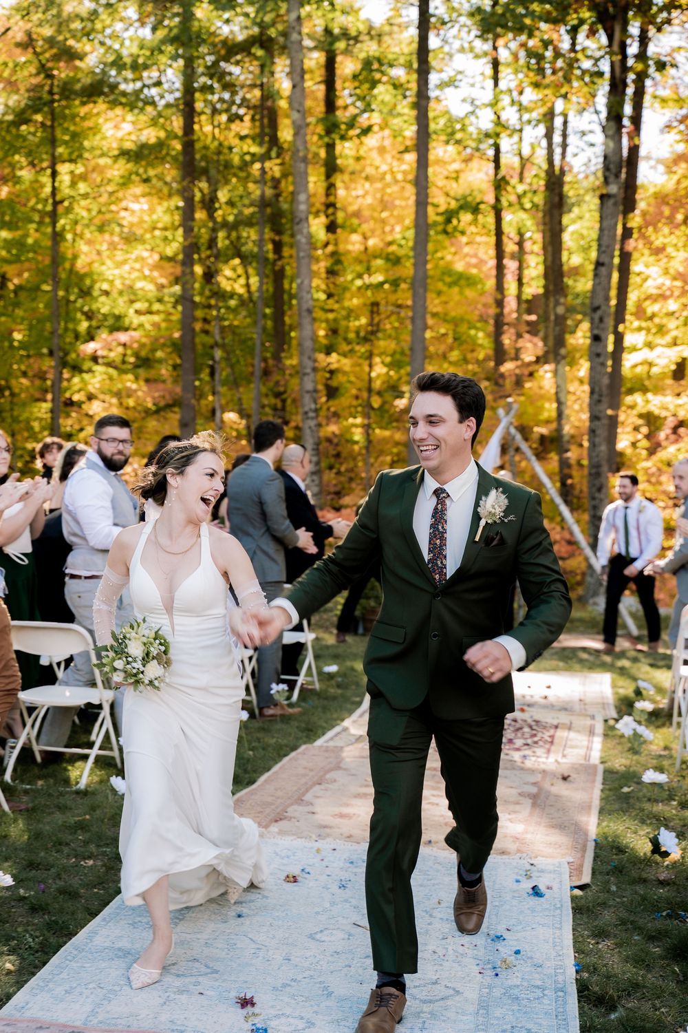 Wedding party members dance joyfully down a white aisle runner in a sunlit autumn forest setting.
