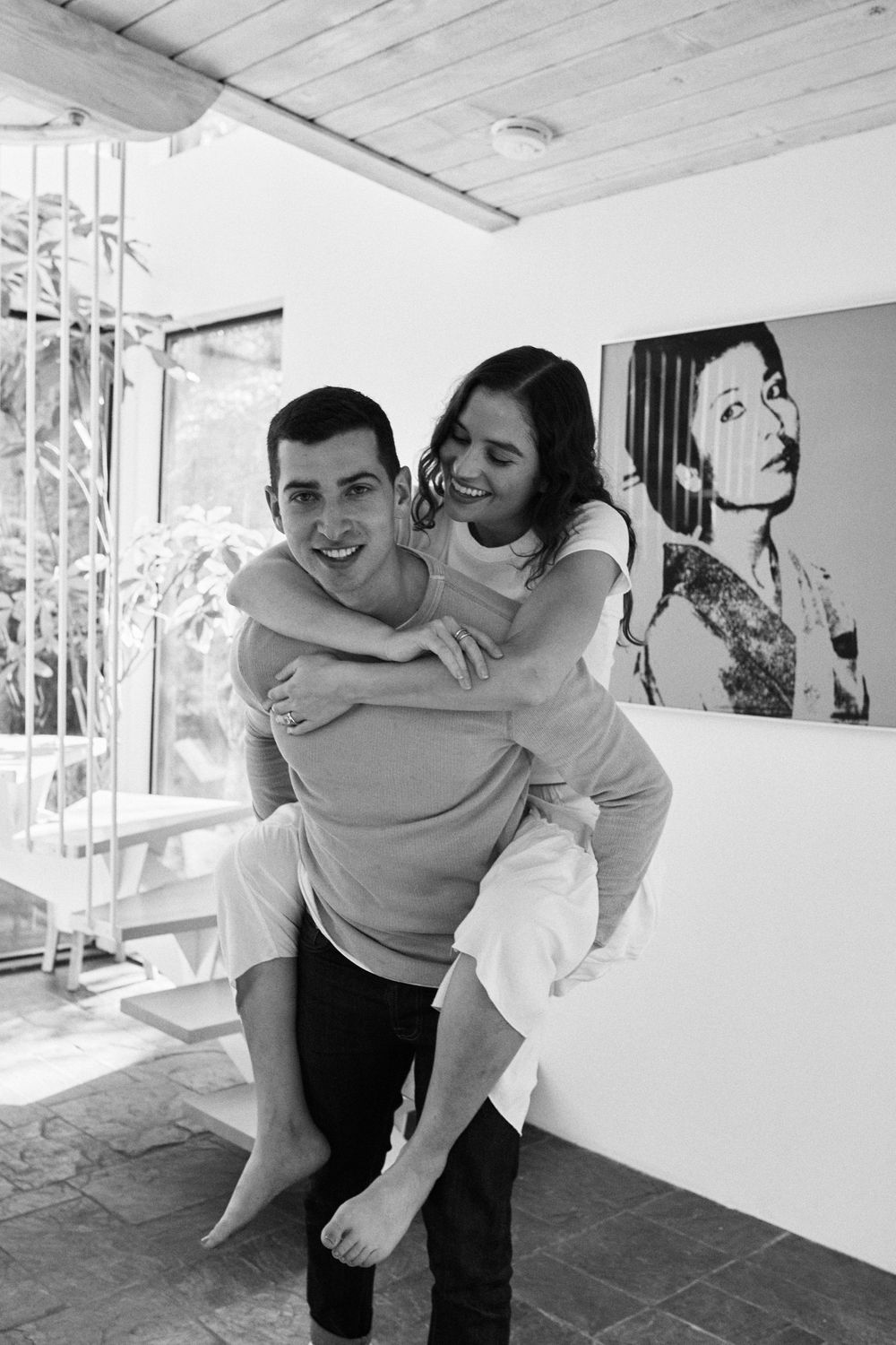 A playful black and white portrait of a couple having fun with a piggyback ride indoors.