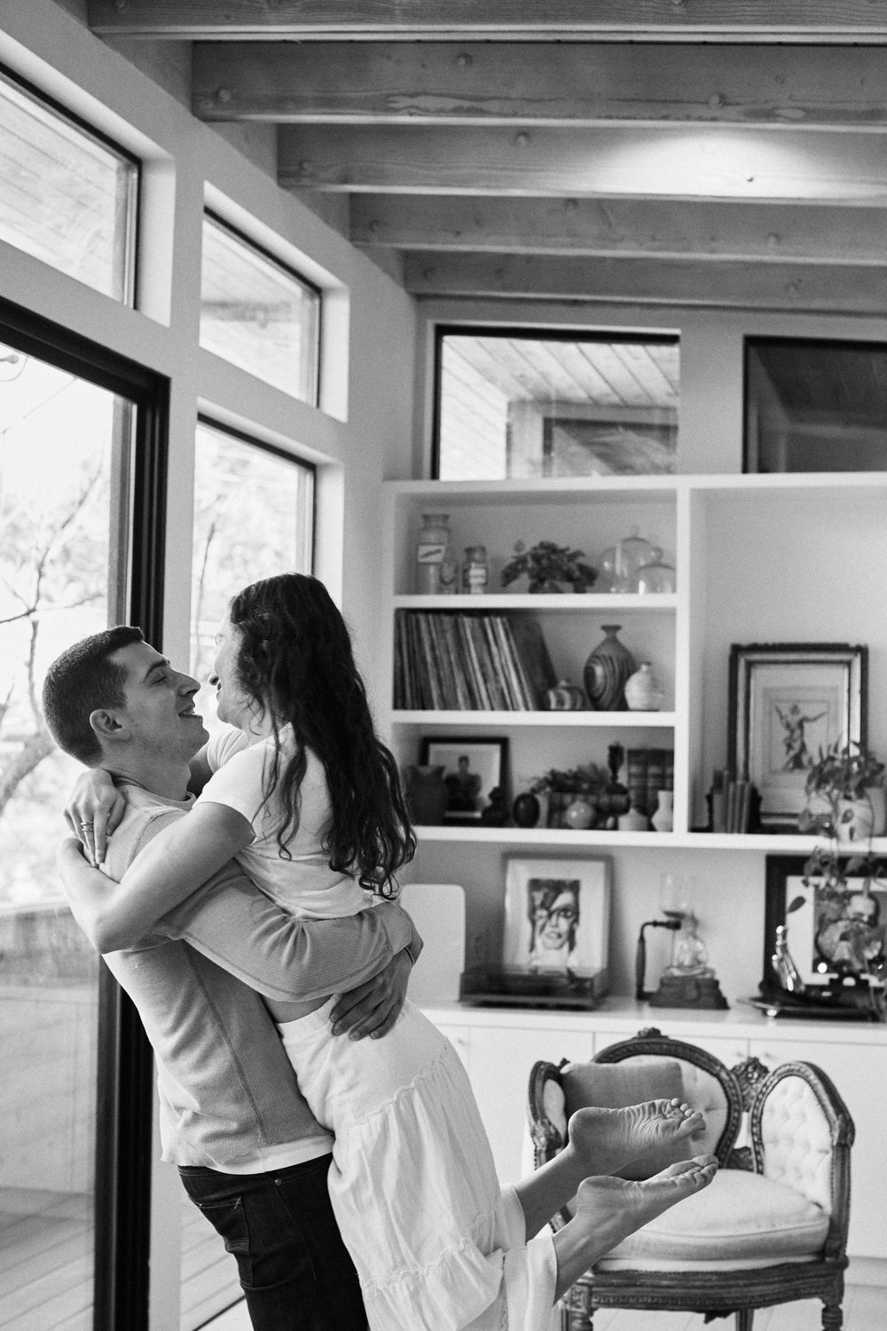 A couple embraces in black and white near a window with built-in shelving and vintage furniture in their home.