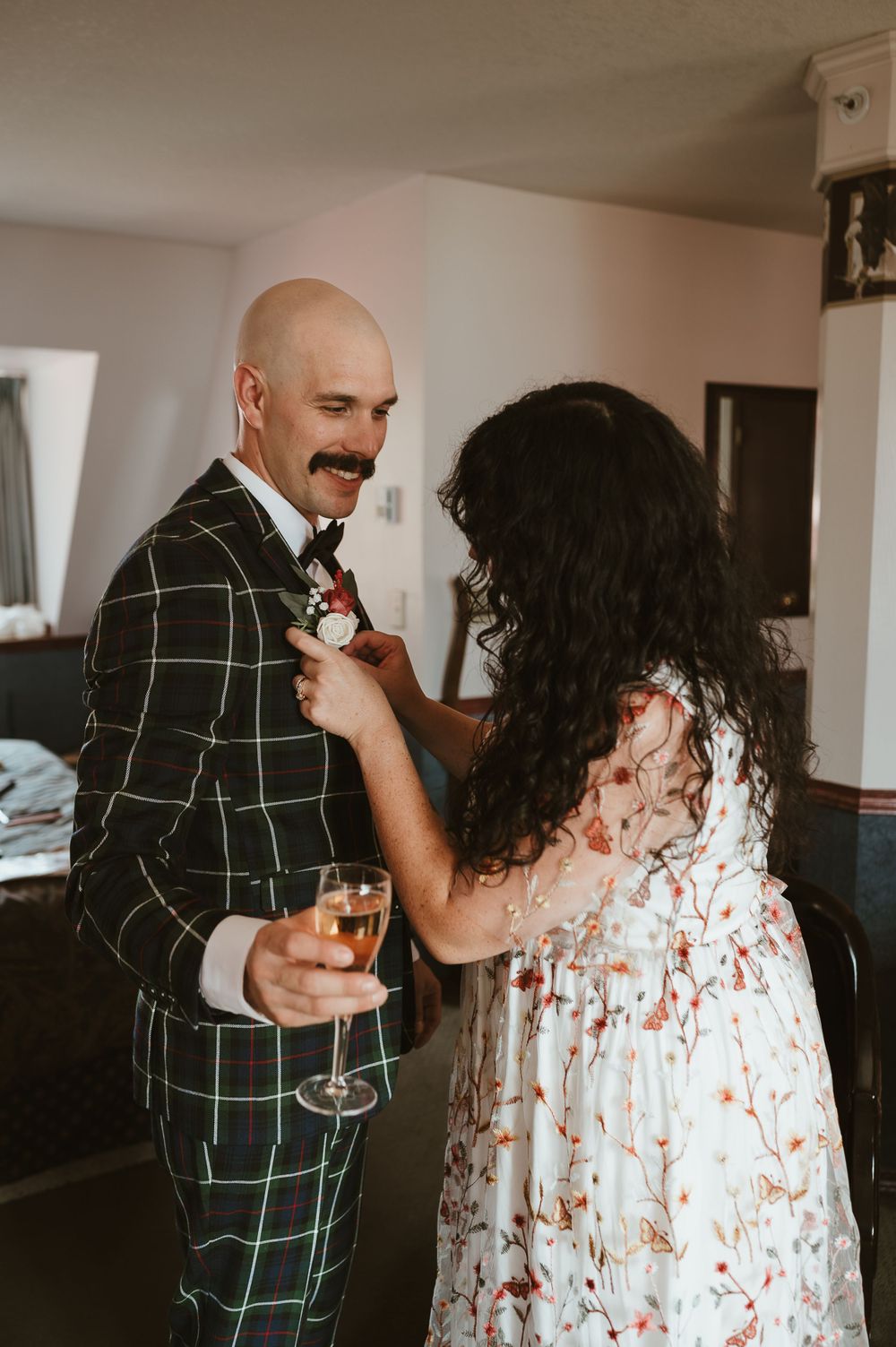 A couple shares an intimate moment as she pins a boutonniere while he holds a champagne glass in a windowlit room.