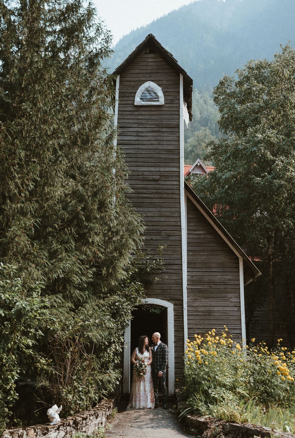 A rustic wooden chapel with tall spire nestled among evergreen trees in a mountain setting.