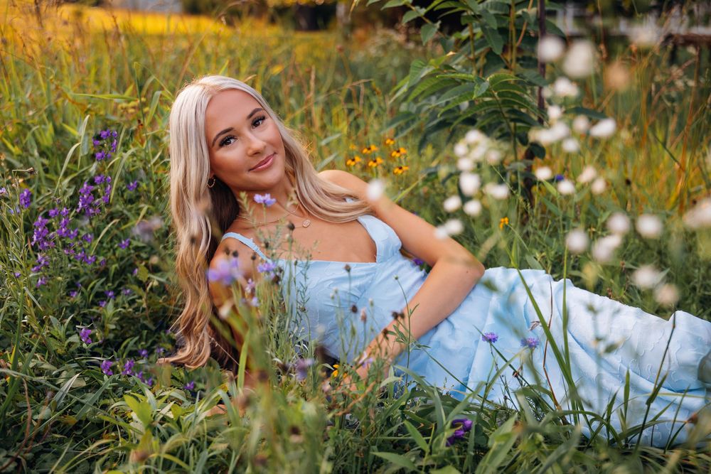 Young blonde woman in a light blue dress lying among wildflowers in a lush green meadow, smiling peacefully.