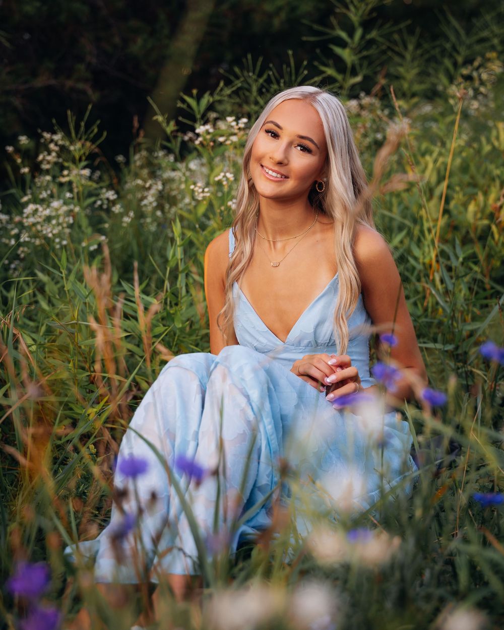 Young woman in light blue dress sitting among wildflowers in a lush green meadow, smiling gracefully.