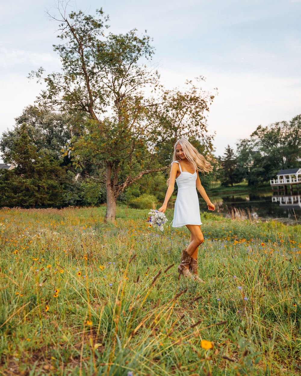 A blonde woman in a white dress and cowboy boots twirls in a wildflower meadow holding a bouquet.
