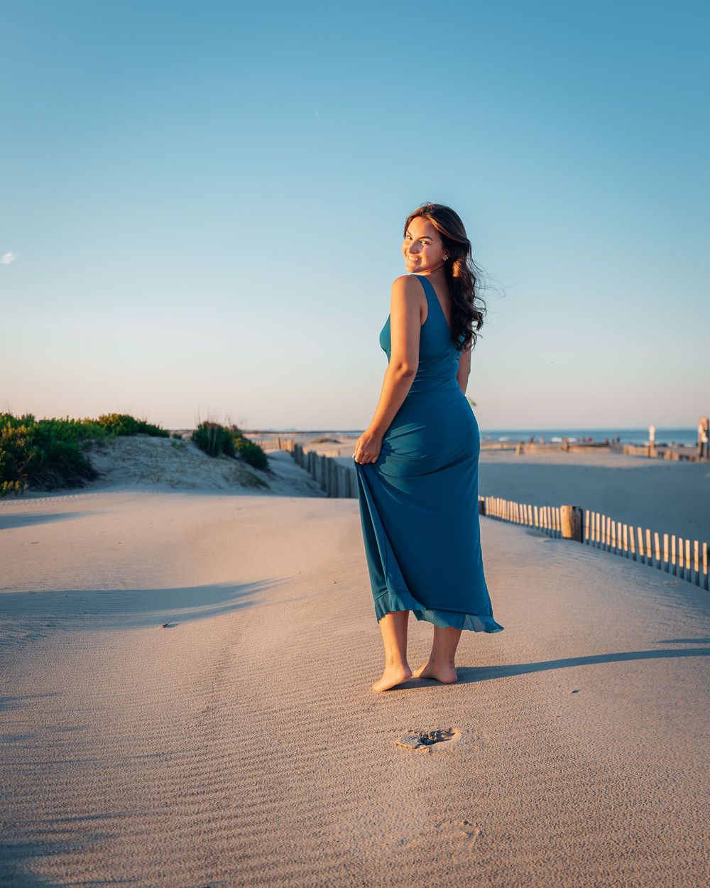 Woman in teal maxi dress standing barefoot on sandy beach at golden hour.