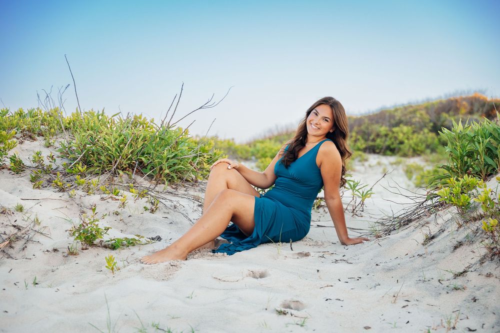A young woman in a teal dress sits gracefully on white sand dunes with green coastal plants under a clear blue sky.