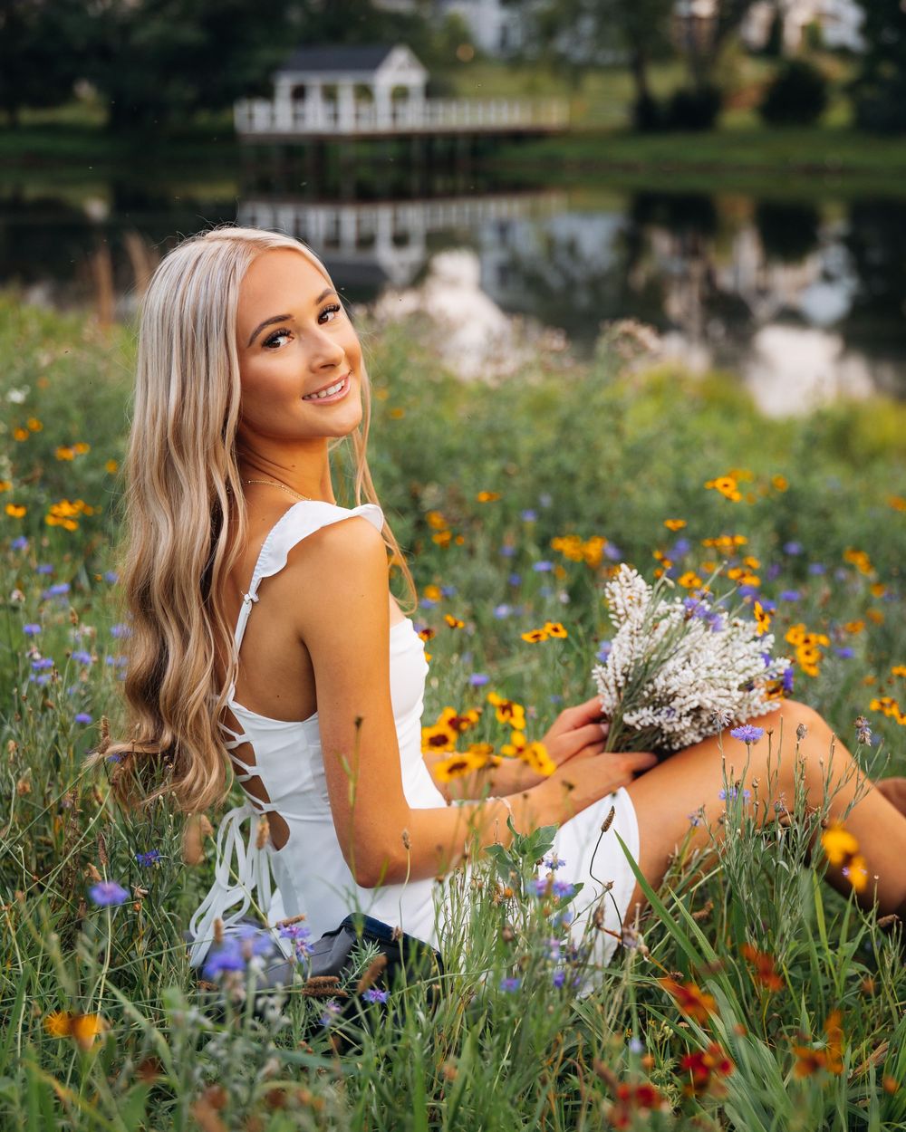 Young blonde woman sitting in a wildflower meadow holding a bouquet, smiling, with a pond and white building behind her.