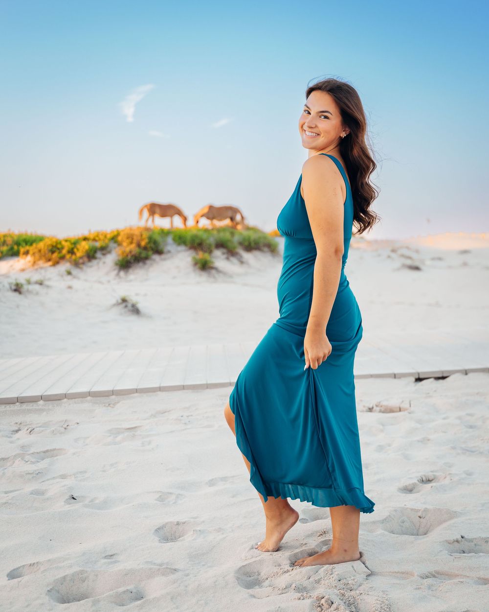 A smiling woman in a teal maxi dress poses on a sandy beach with wild horses grazing on dunes behind her.